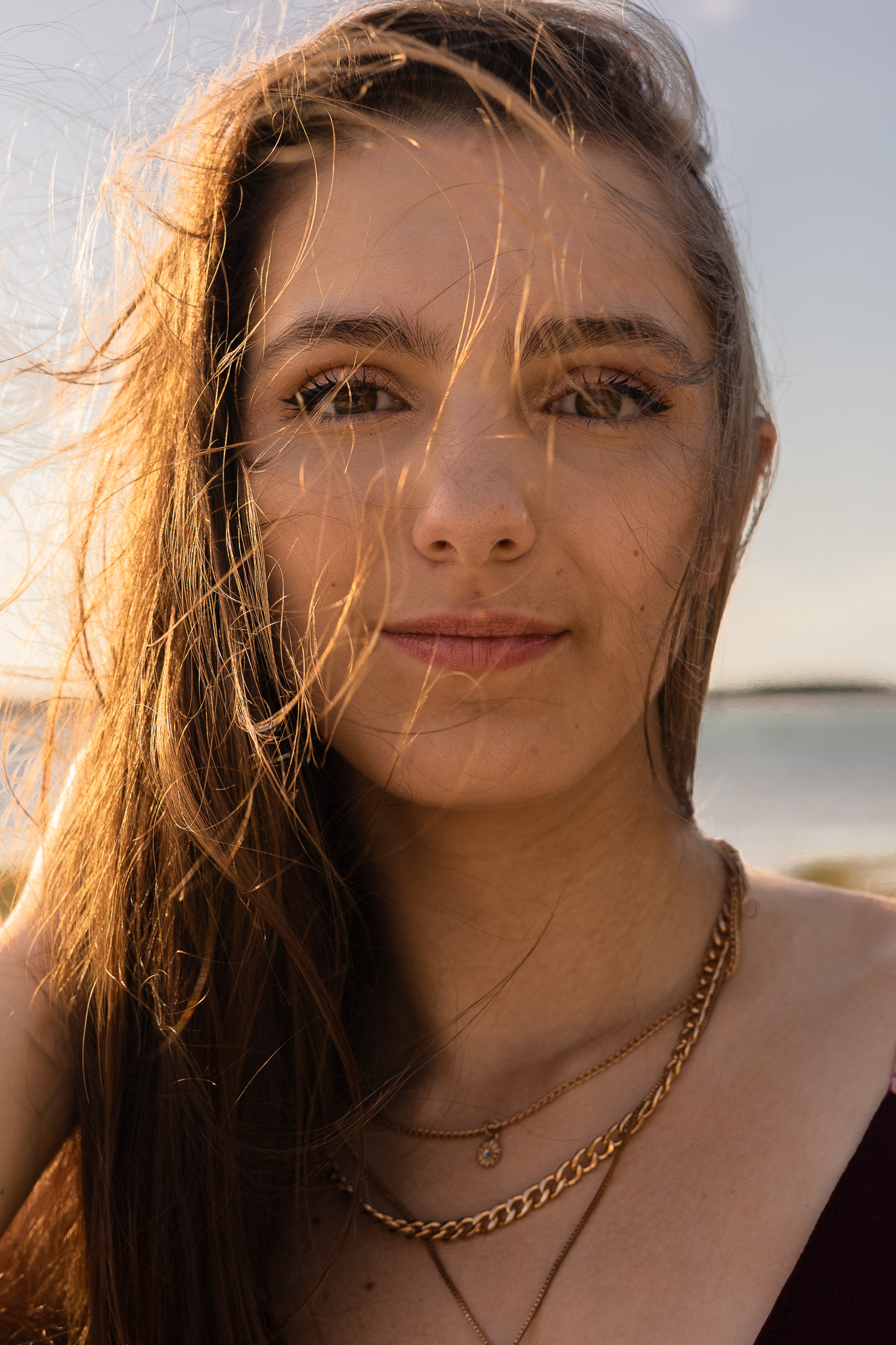 Senior portrait of a young woman with windswept hair and gold layered necklaces at golden hour on a Florida beach.