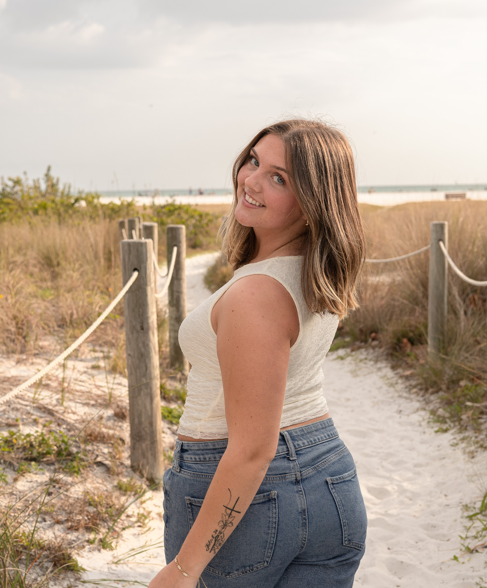 Senior portrait of a smiling young woman in a white tank and jeans on a sand path at a St. Pete Gulf beach.