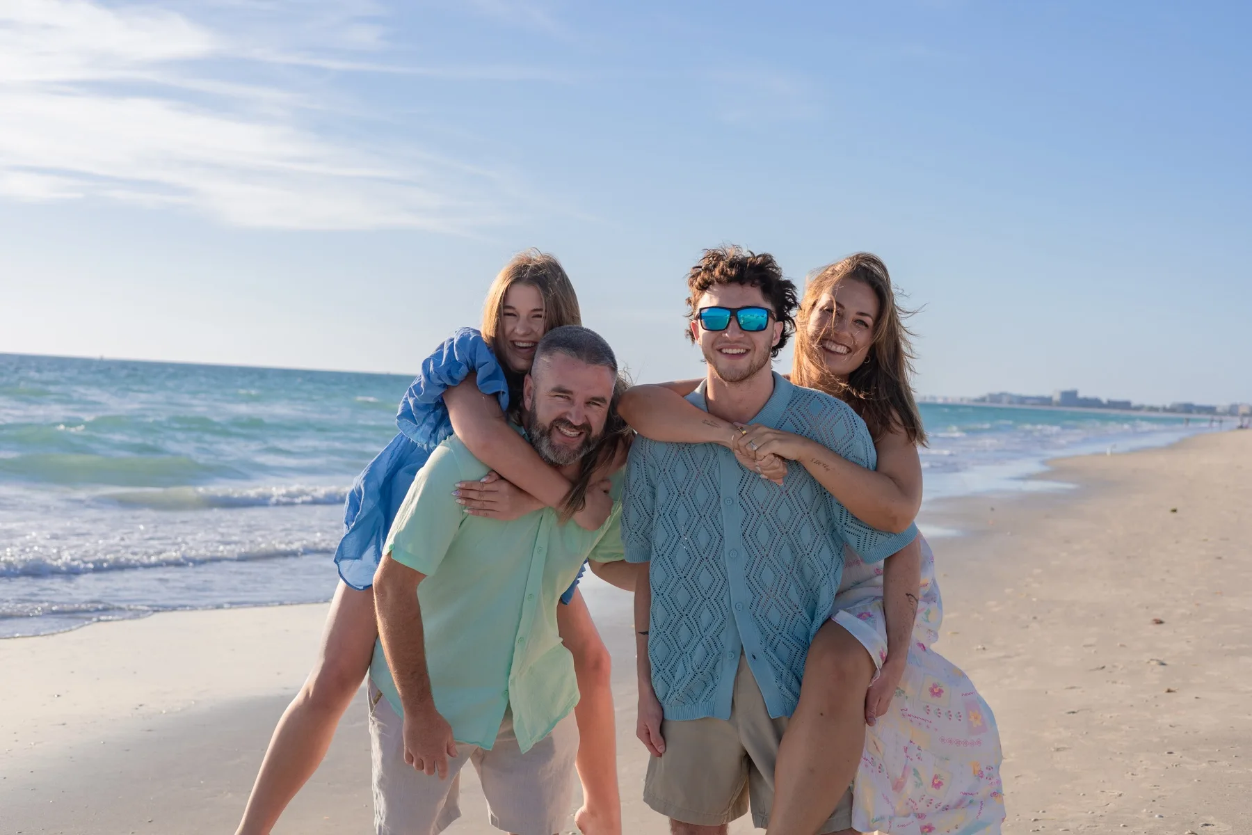 Two couples piggyback, laughing along the shoreline.