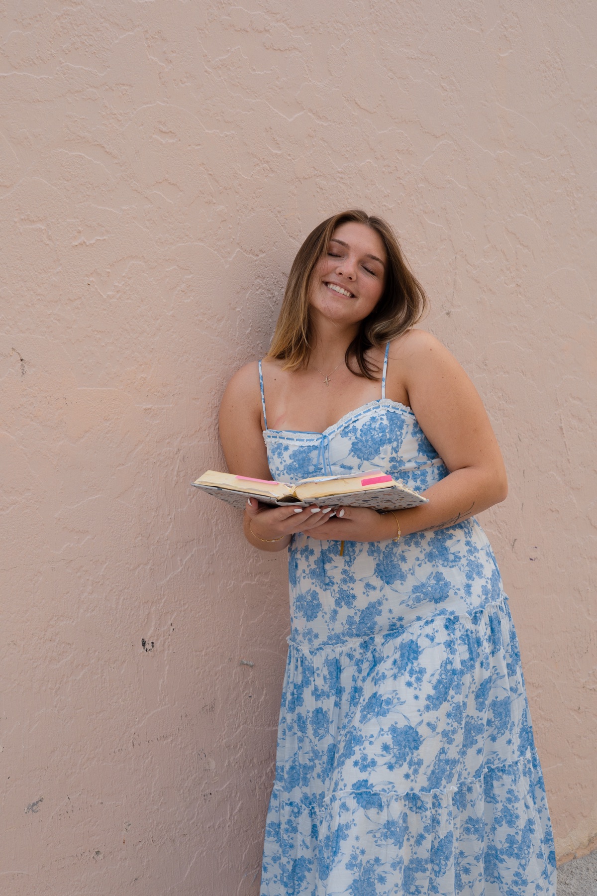 Eyes-closed smile against a soft pink wall, book in hand