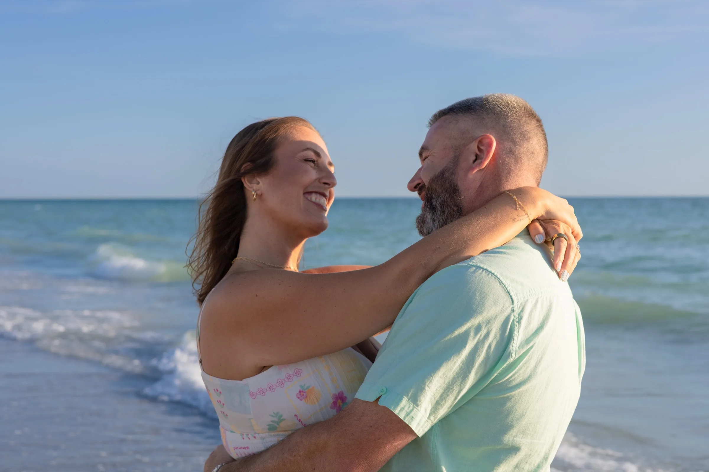 Couple in a soft embrace on a Florida Gulf beach at golden hour, smiling at each other against the open horizon.