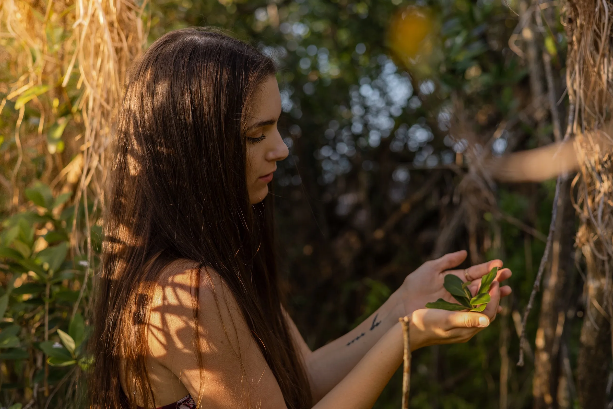 Creative portrait of a young woman holding a green leaf in dappled golden light among hanging vines and tropical foliage.